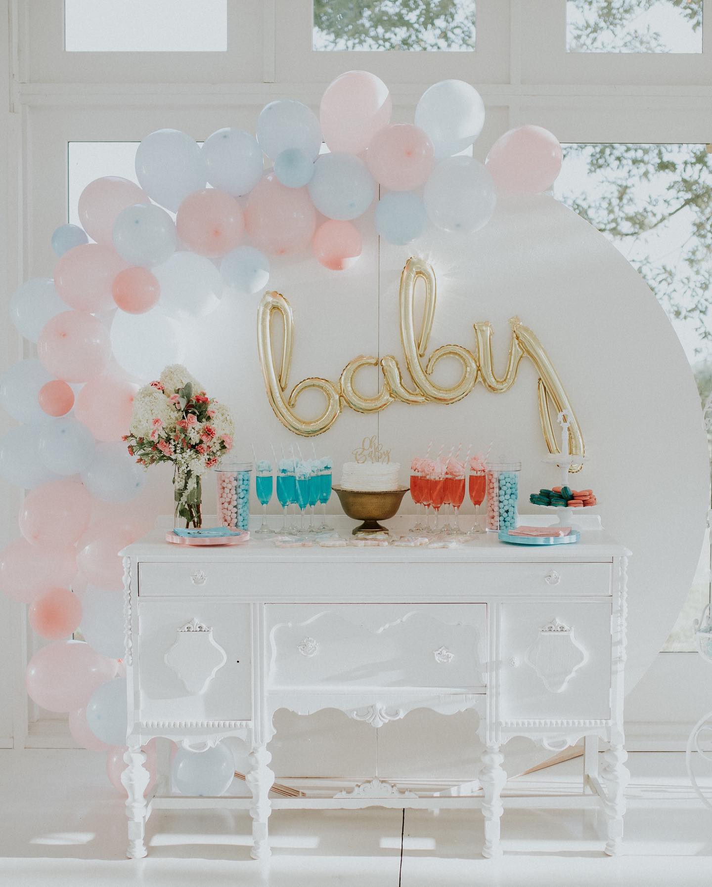 baby shower balloon arch with pink and blue drinks and cake on a white table